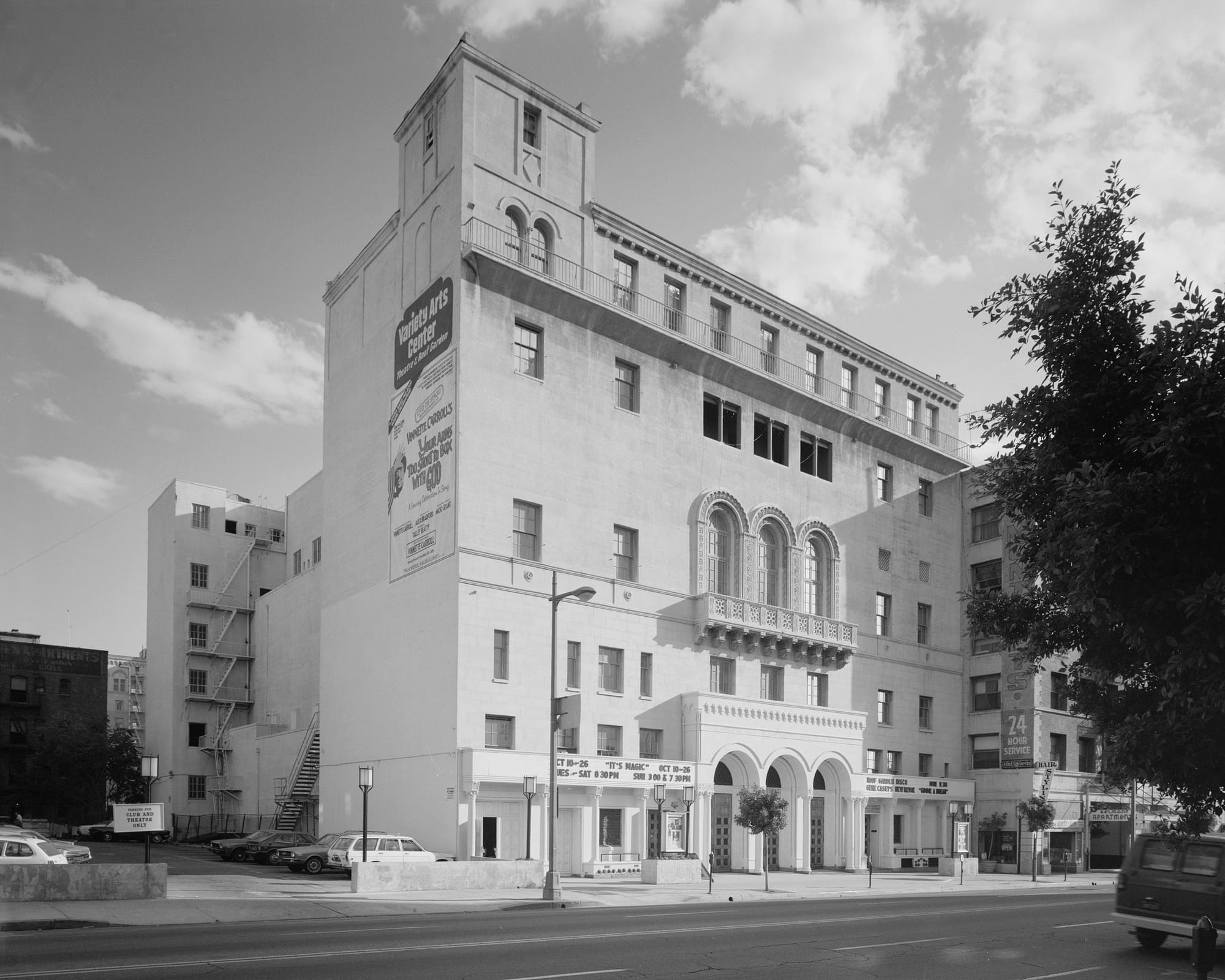A black and white image of a classical theater building with large arch windows, on a busy city street.