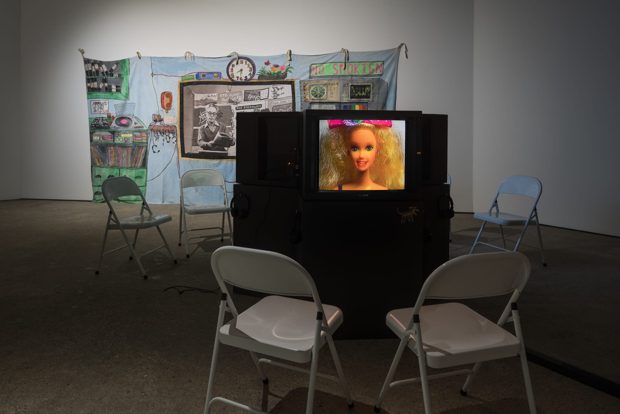 Gallery view of a monitor showing a barbie doll with two folding chairs placed in front of it, and in the background on the gallery wall is a colorful collage tapestry.