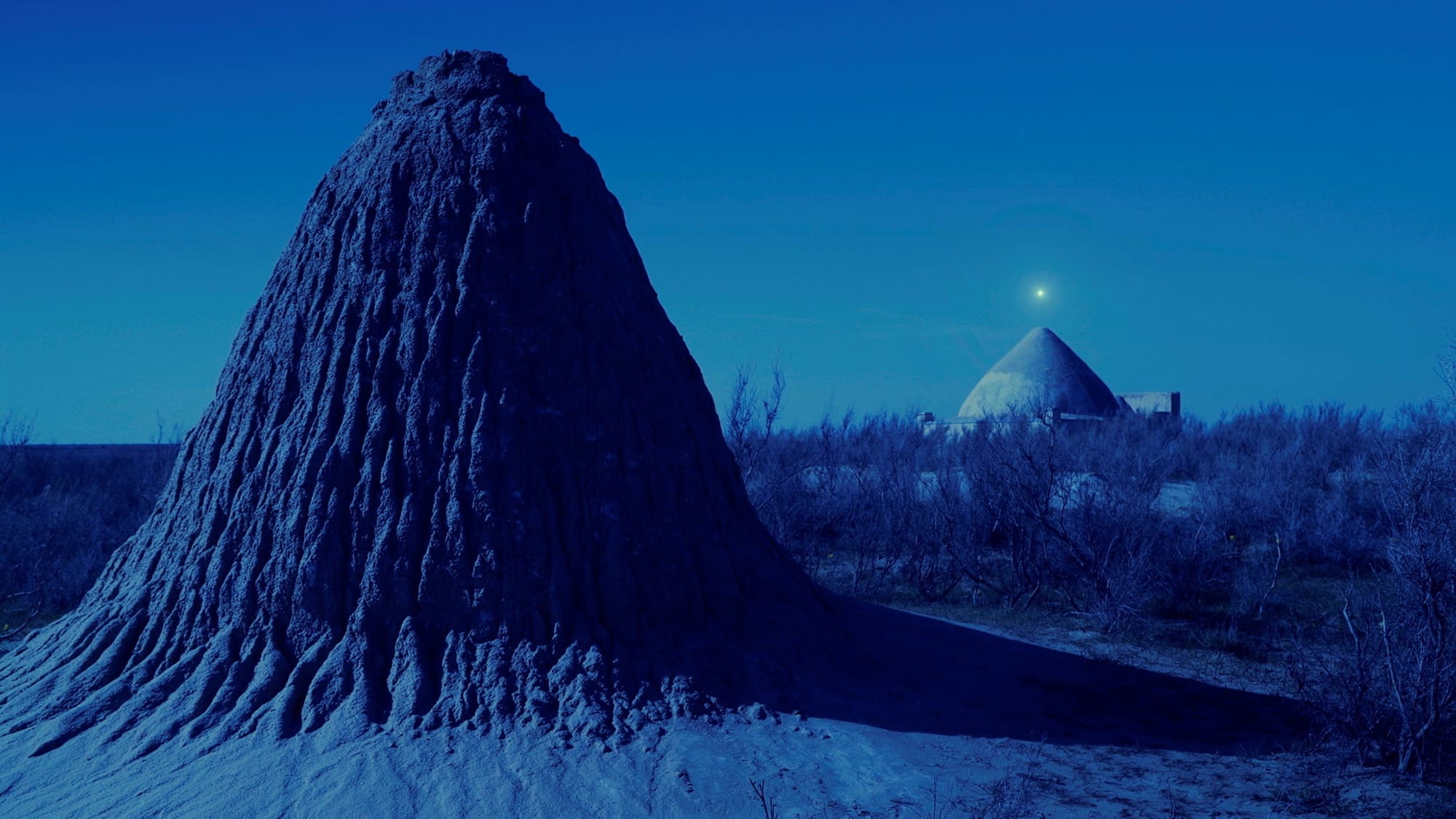 A blue-tinted image of a small hill in the foregroud and a built structure in the backgroud with a star shining above it.