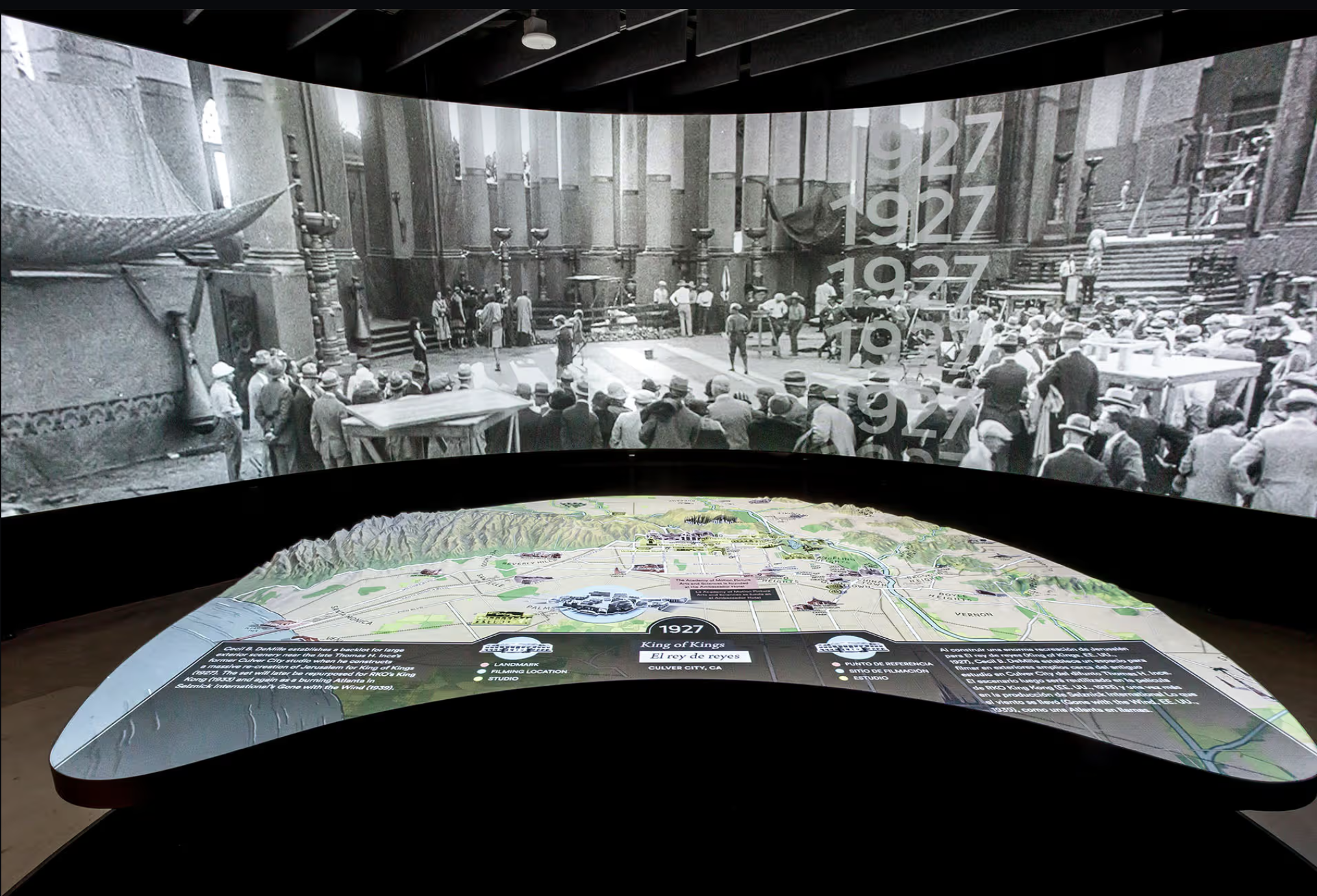 An installation with a large screen depicting black and white images of an old film set with an interactive display below it that diagrams the cultural geography of Hollywood.