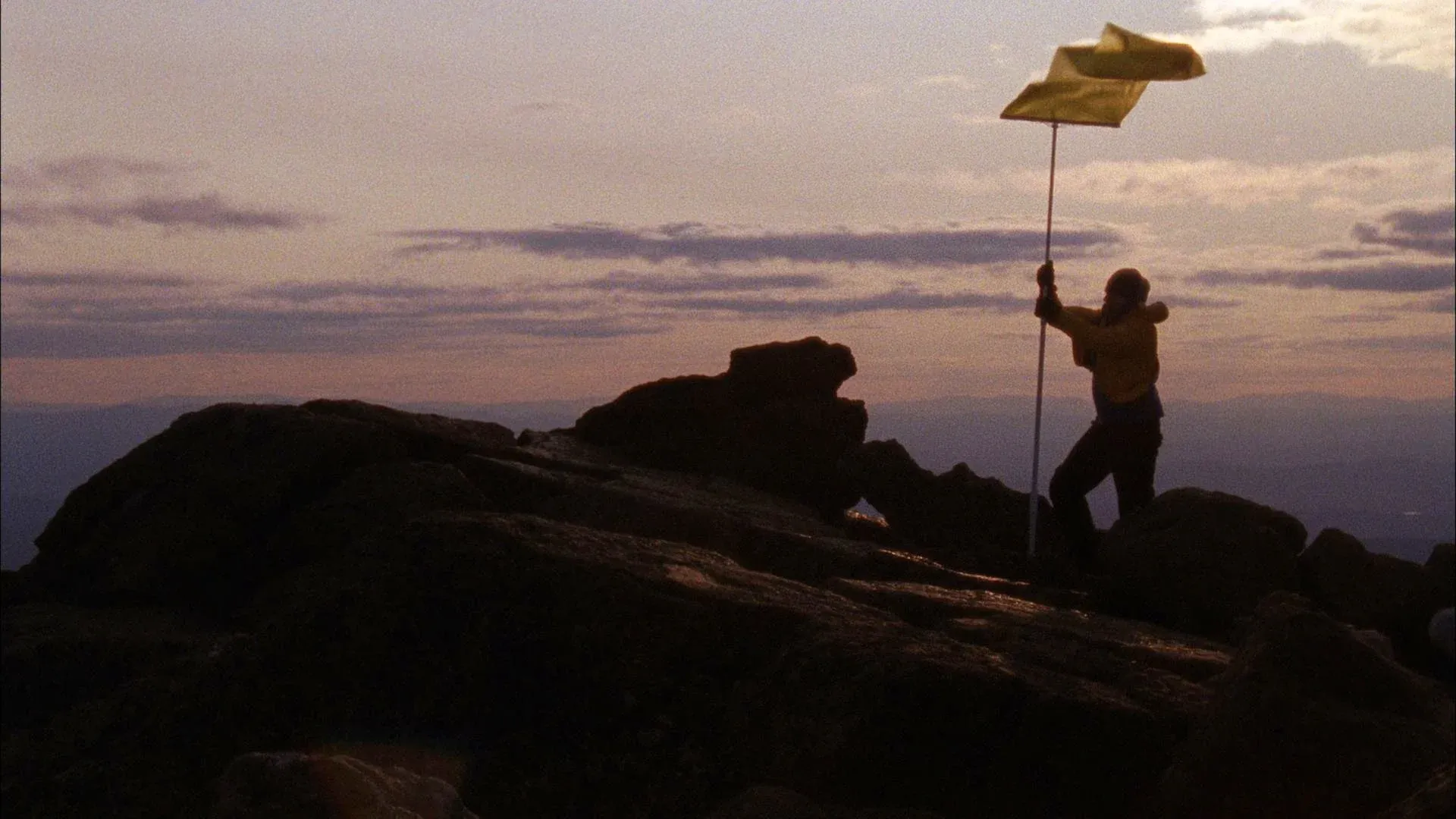 A figure holding a flag, standing on rocky terrain, is seen in silhouette at dusk.