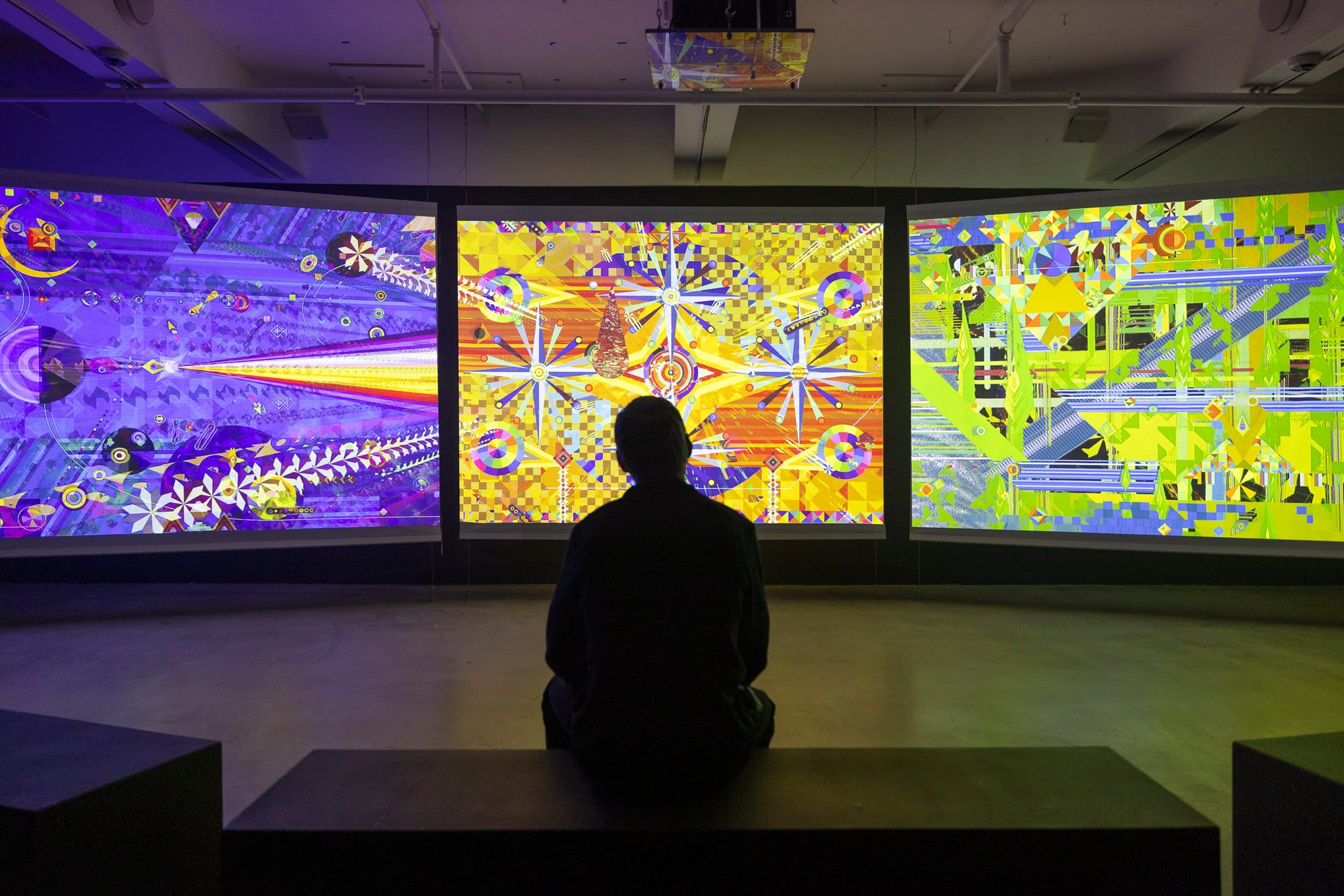 A figure in silhouette sits on a bench in a dark gallery in front of three screens projecting brightly-colored and abstract geometric animation.