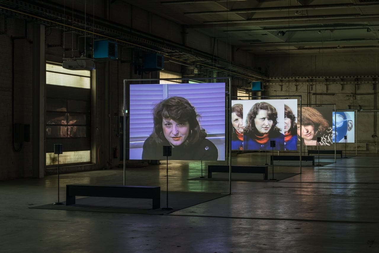 An expansive gallery space with multiple large screens, each with the same woman's face speaking in a direct camera address.