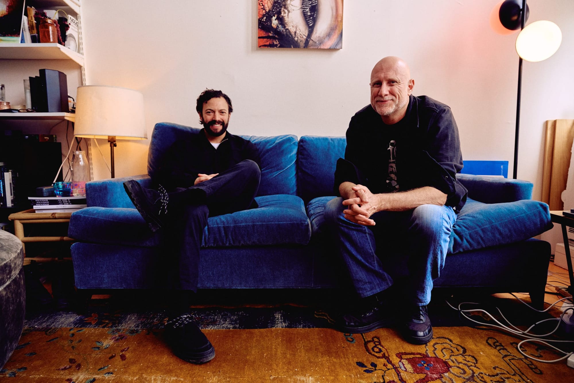 Two smiling men dressed in dark colors, sitting on a blue couch with lamps on either side and a bookshelf next to it.
