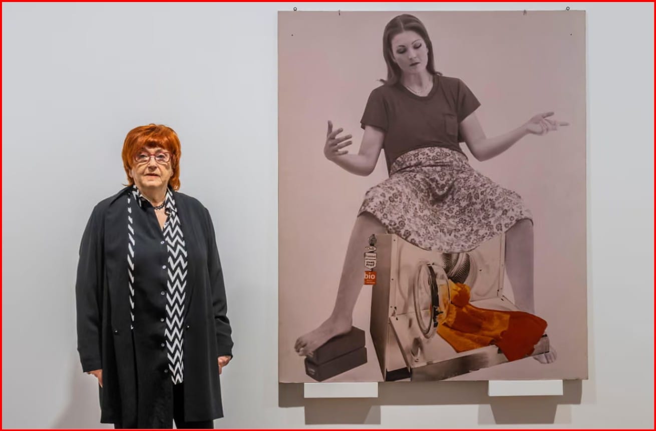 An older woman with red hair and glasses, wearing black clothes with a black and white scarf, standing next to a large collage featuring a woman with her legs spread, giving birth to a washing machine.