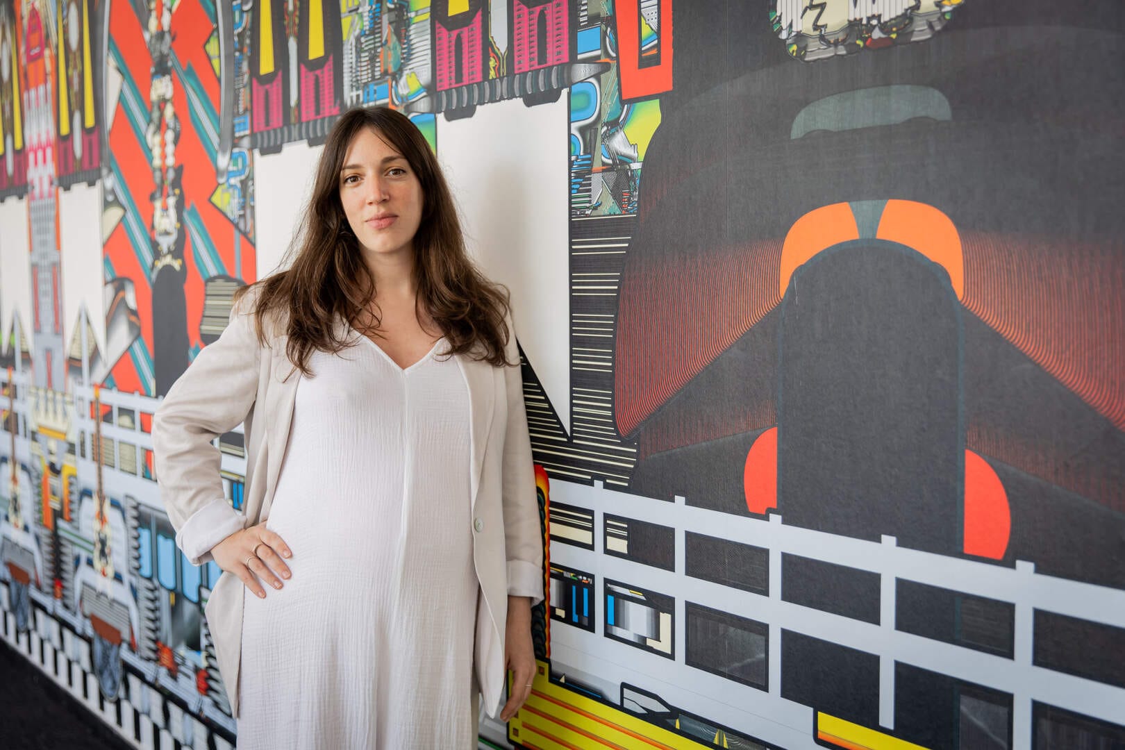 A woman with long brown hair, wearing a flowing white dress, stands in front of a colorful abstract mural.