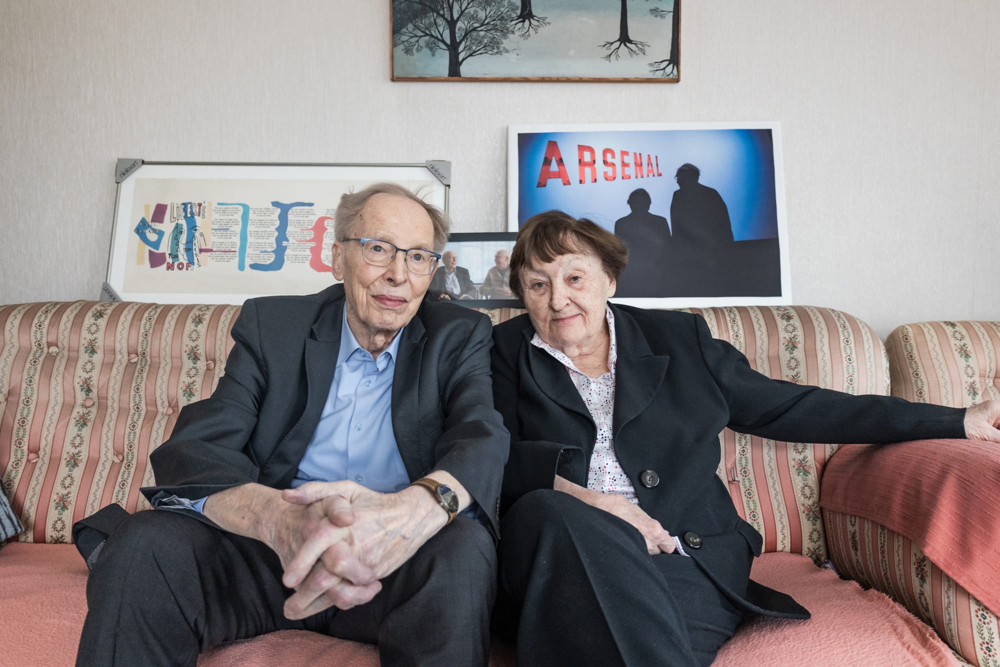 An elderly white couple dressed in black formal attire, sitting on a couch with framed photos behind them, one of which is a picture of the two of them in silhouette standing in front of a sign that reads Arsenal. 