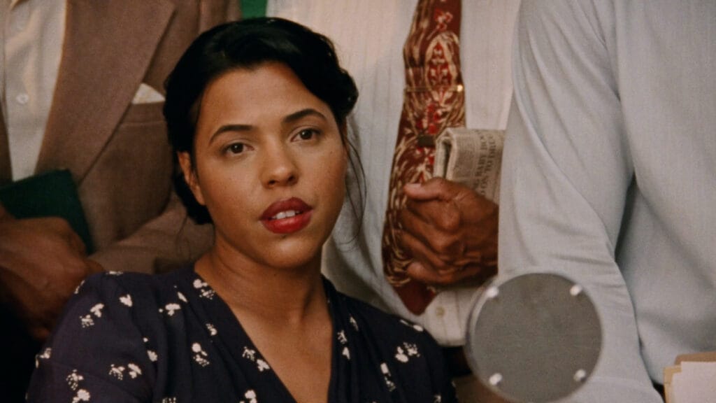 A Black woman with short hair wearing a dark blue dress, sitting down with men in suits standing behind her.