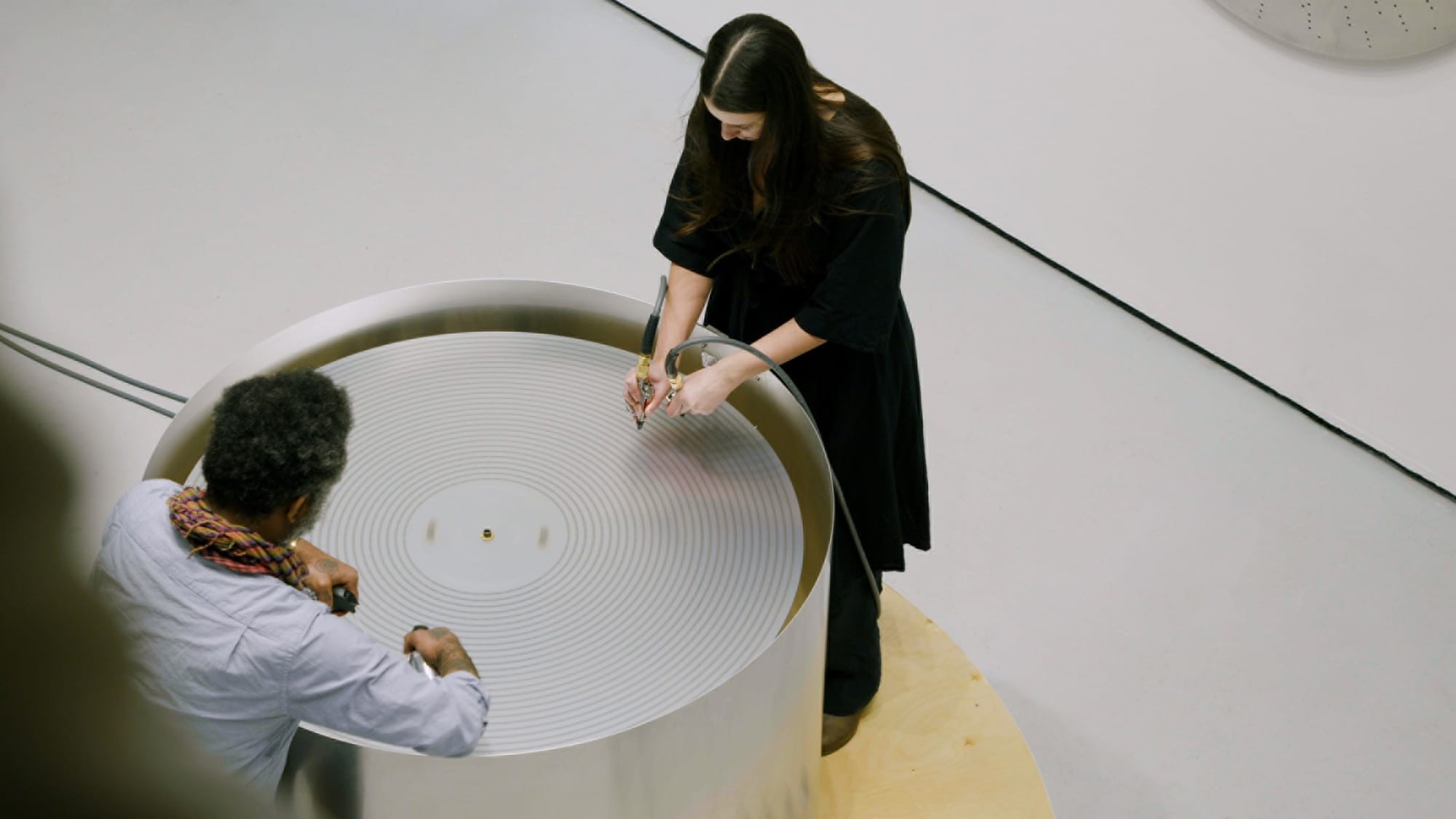 A man and a woman stand over a circular tub with concentric cirlces as grooves on it, with the woman holding an instrument that reads the grooves.