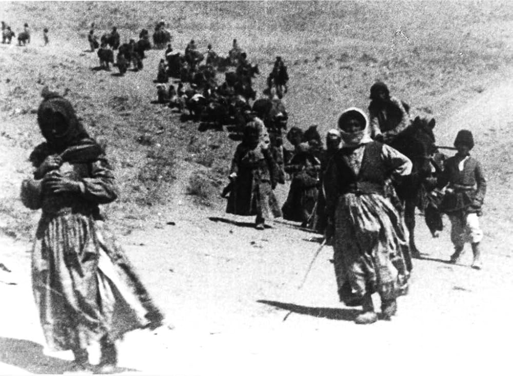 A grainy, black and white archival film image of early twentieth century migrants traveling in a column on a dirt road.