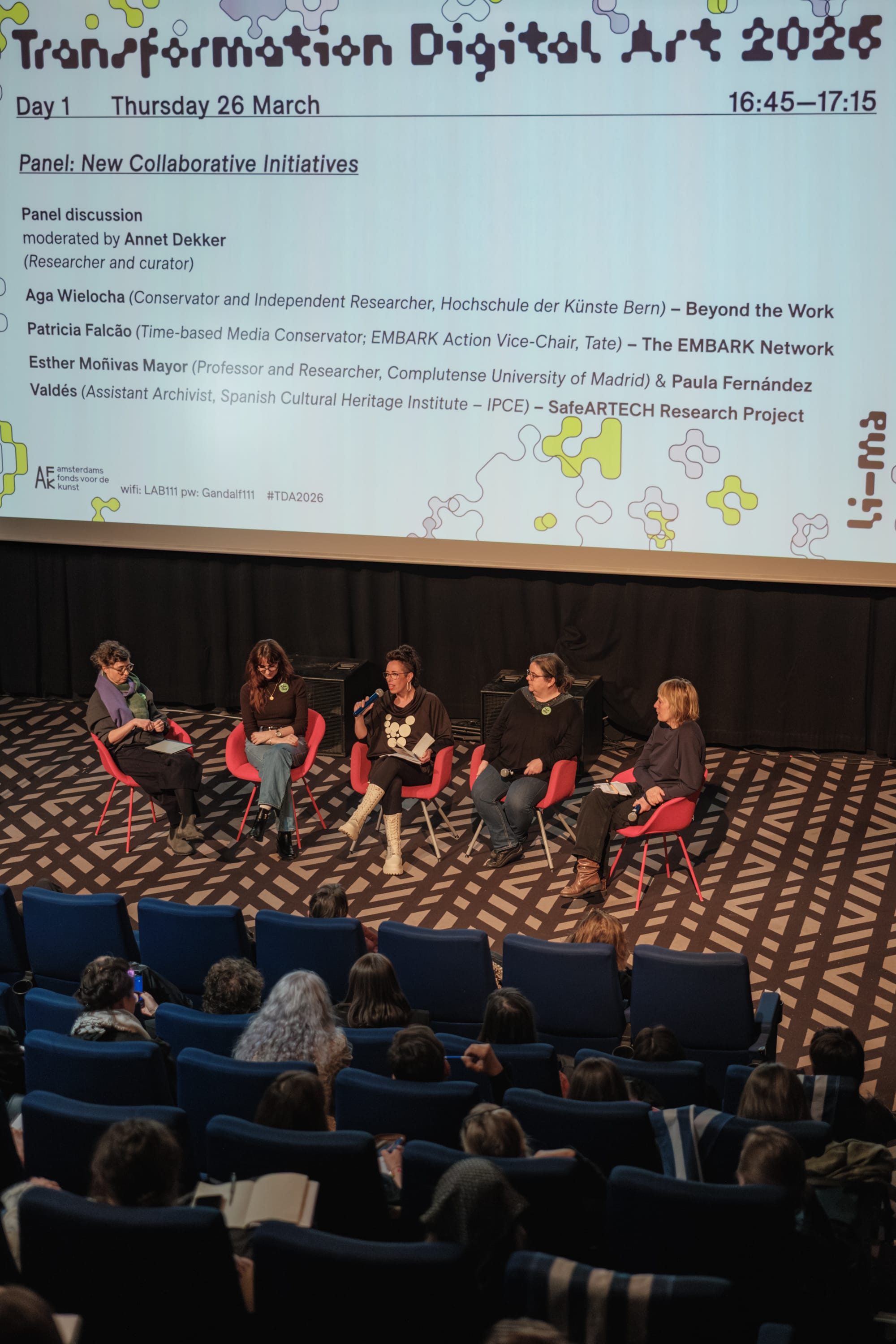 Five panelists at a conference sitting under a large video screen that details their names and titles.