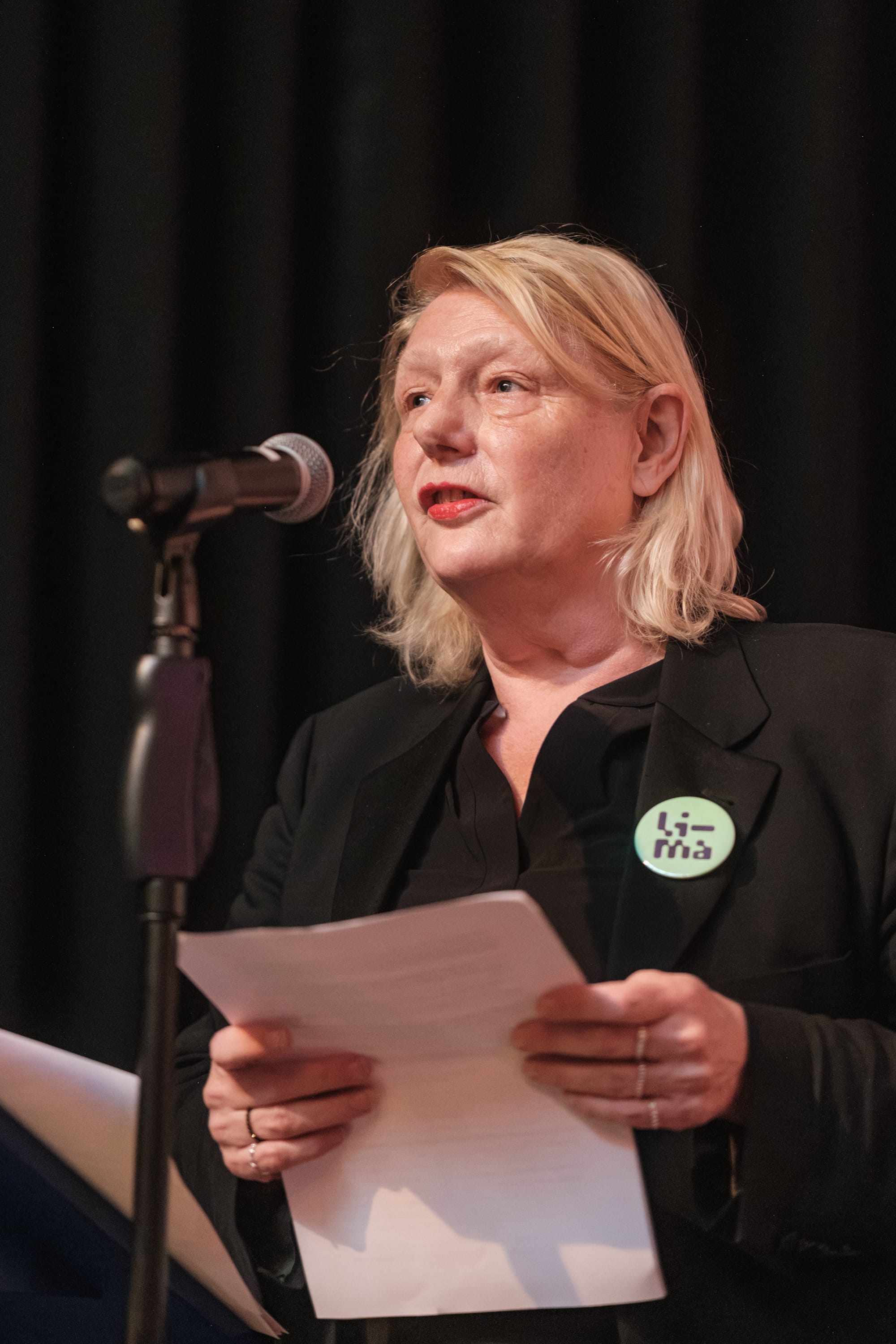A woman with black clothes and blonde hair, speaking into a microphone and holding papers, wearing a pin on her jacket lapel that reads LI-MA.