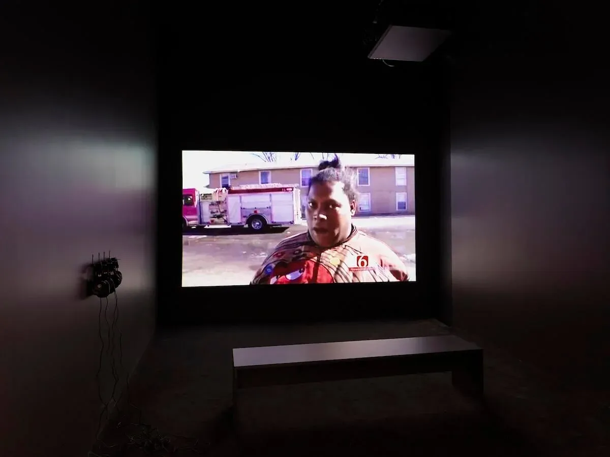 A dark gallery space with a large video projection of a Black woman standing in front of a fire engine.