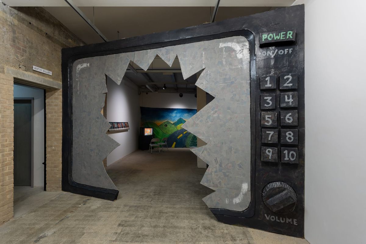 A gallery view of an immense television with a broken screen that visitors walk through to access another gallery with monitors and tapestries.