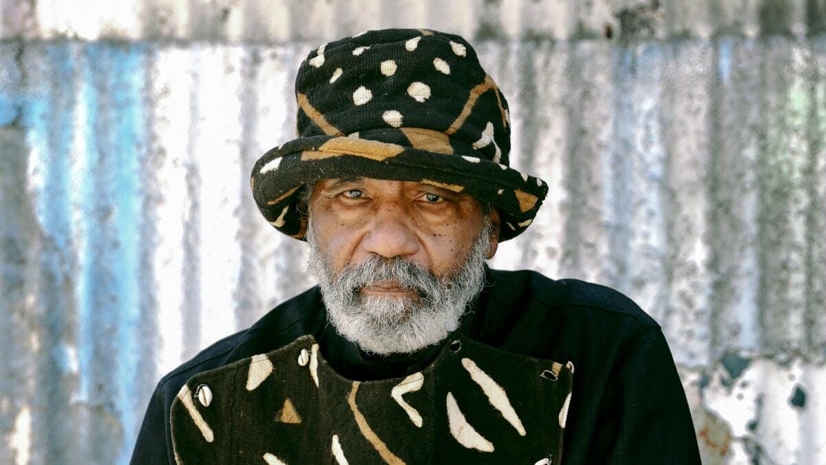 An elderly Black man with a full gray beard wearing a matching hat and sweater with beige and white patterns.