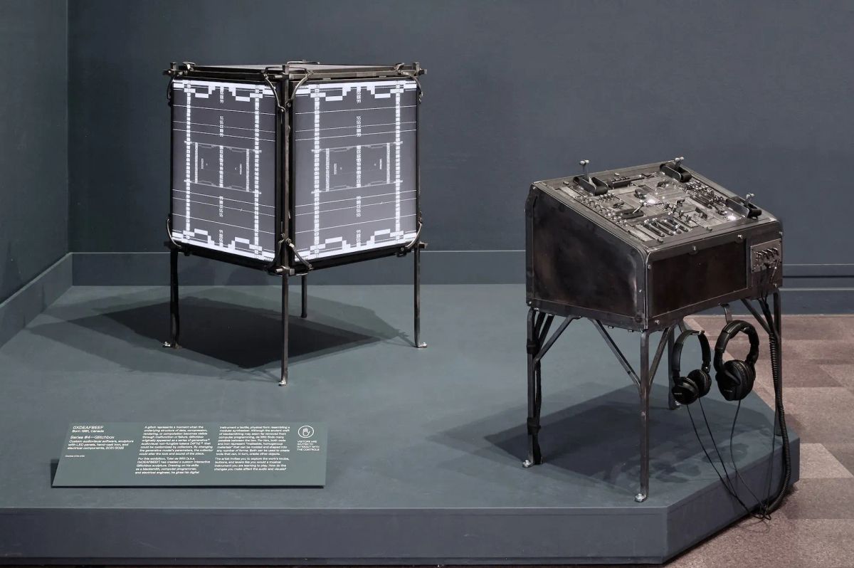 A four-sided screen device showing digital art on a gallery platform with a control panel next to it with various switches, levers and headphones.