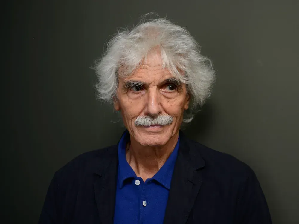A smiling man with long gray hair and a thick gray mustache, wearing a blue collared shirt with a dark jacket over it, standing against a neutral backdrop..