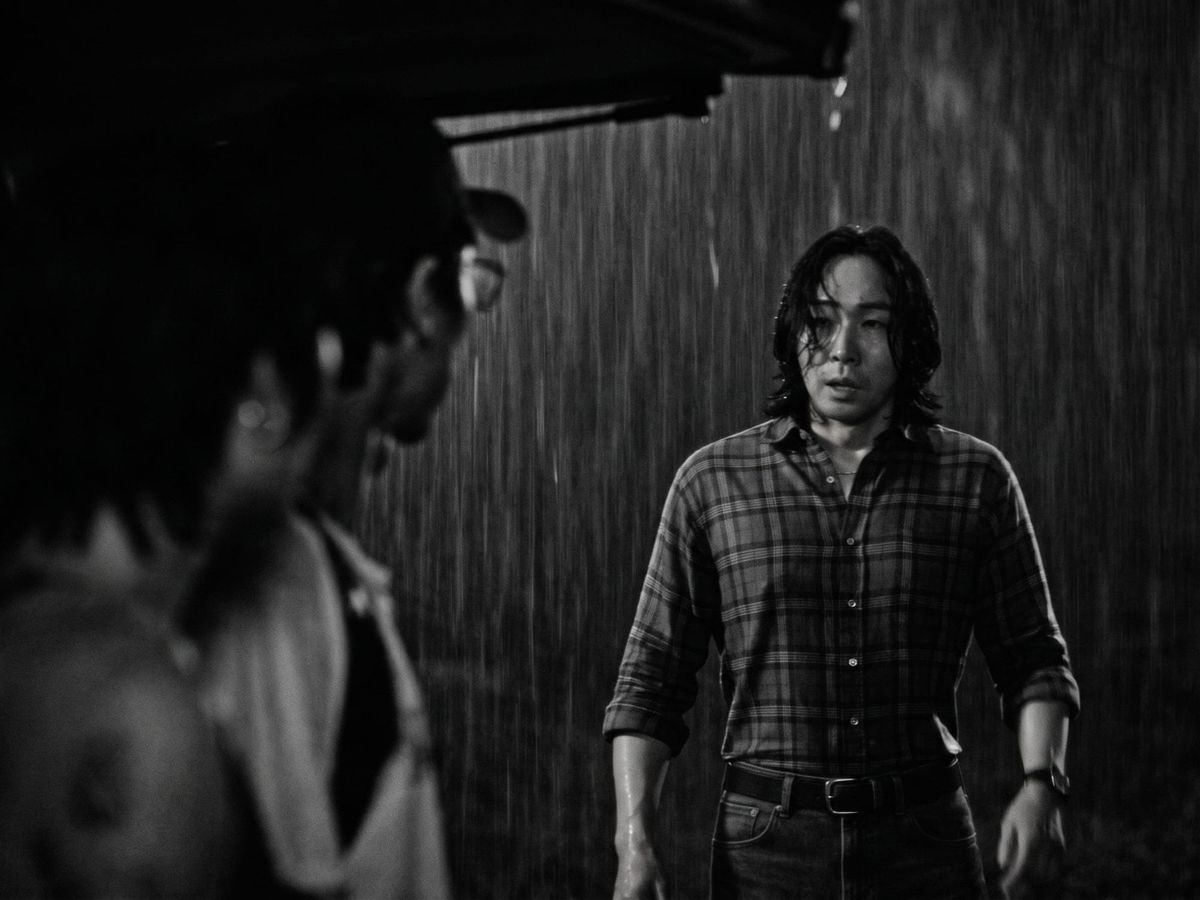 A black and white image of a man with long hair wearing a plaid shirt and standing in the rain.