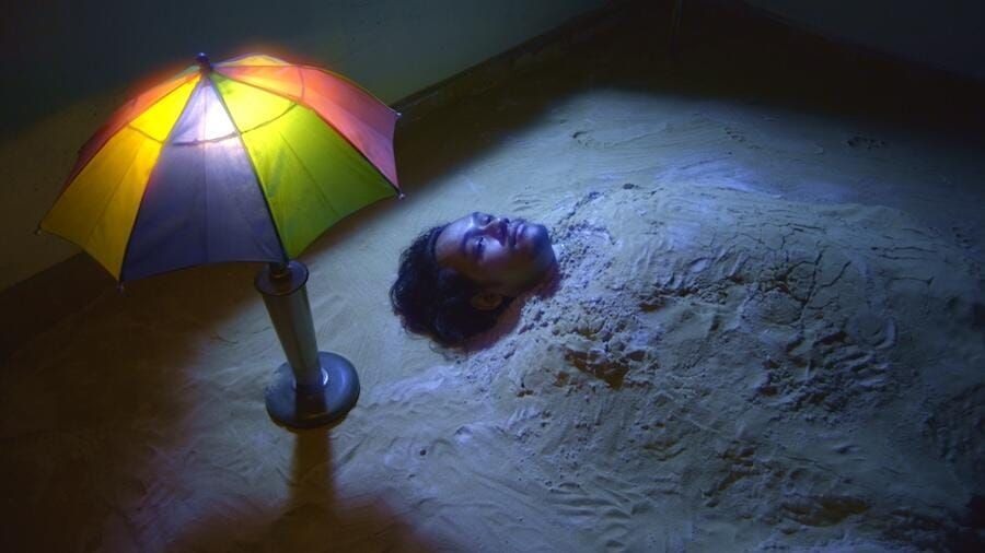 An individual buried in sand all the way up to their neck, with a lamp in the shape of an umbrella next to them.