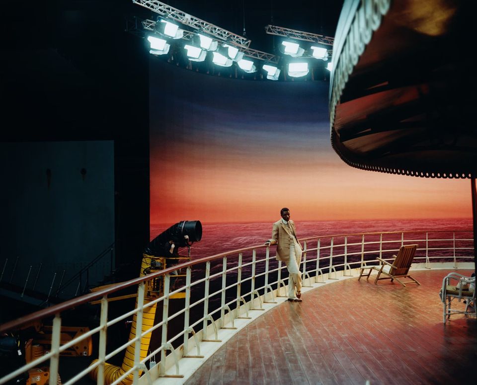 A movie set with a Black man in a beige suit standing on the deck of an ocean liner, with lights above him and a painted backdrop of the sea at sunset behind him.