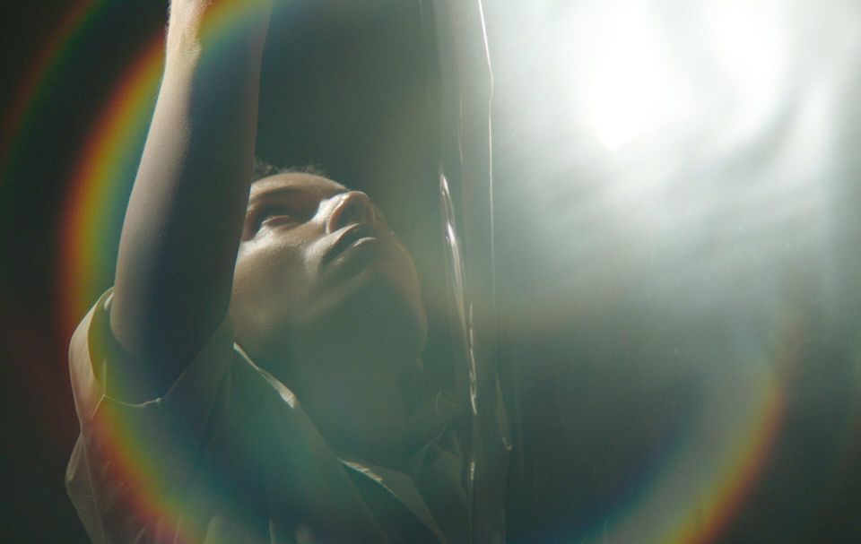 A woman wearing a white shirt and with her arms raised looks up, with direct light creating lens flares around her.
