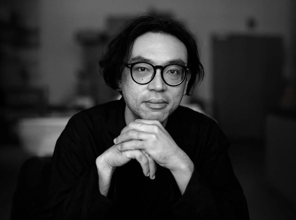 A black and white photo of an Asian man with medium-length hair, dressed in black, wearing glasses, with his hands folded, positioned against an out of focus background.