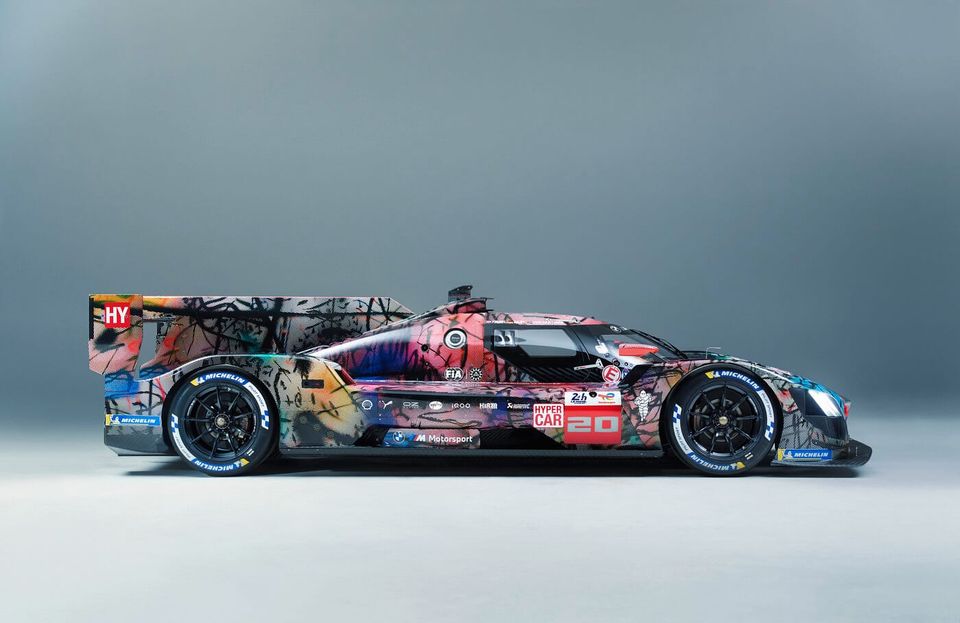 A racing car hand painted in multiple colors, with stickers and other artistic markings on it, positioned against a flat off-white backdrop.