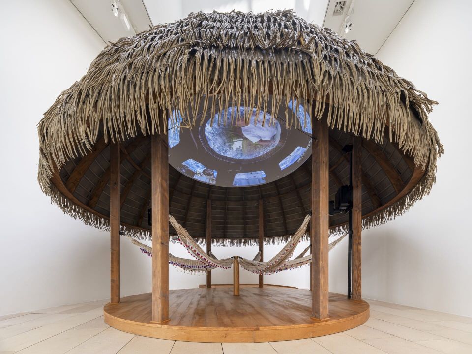 An African hut placed inside of an art gallery, with video images projected onto the roof. 