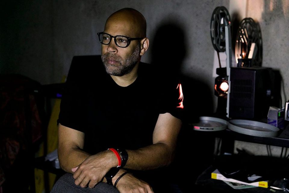 A Black man with glasses in a black t-shirt sitting in front of a 16mm projector.