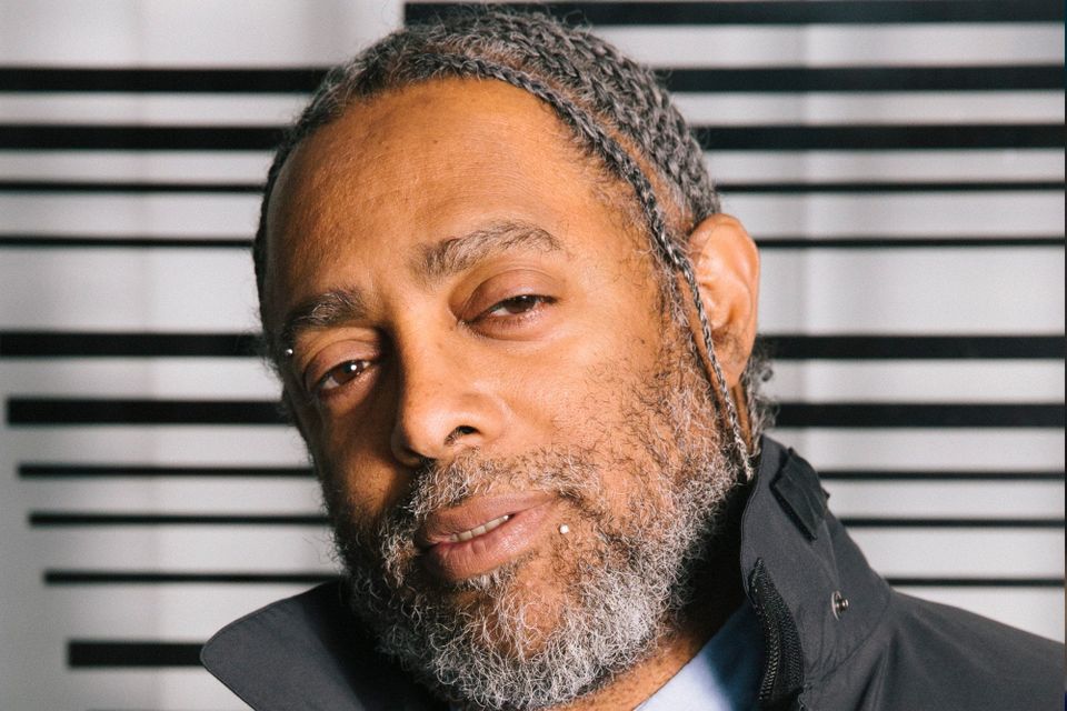 A older black man with braids and a gray beard, wearing a dark jacket, standing in front of a white background with black stripes.