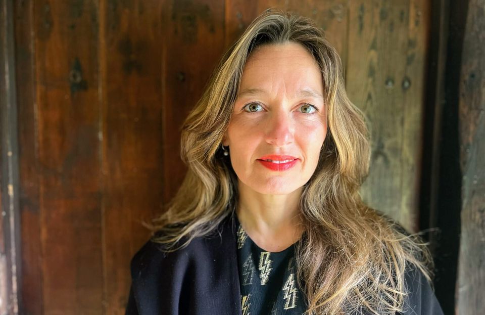 Portrait of a woman with long blonde hair and green eyes, wearing a black pullover, standing in front of a wooden wall.