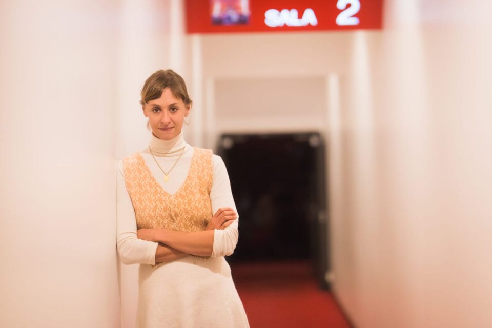 A woman with brown hair wearing a white dress and a brown vest, leaning on a wall in the hallway of a cinema.