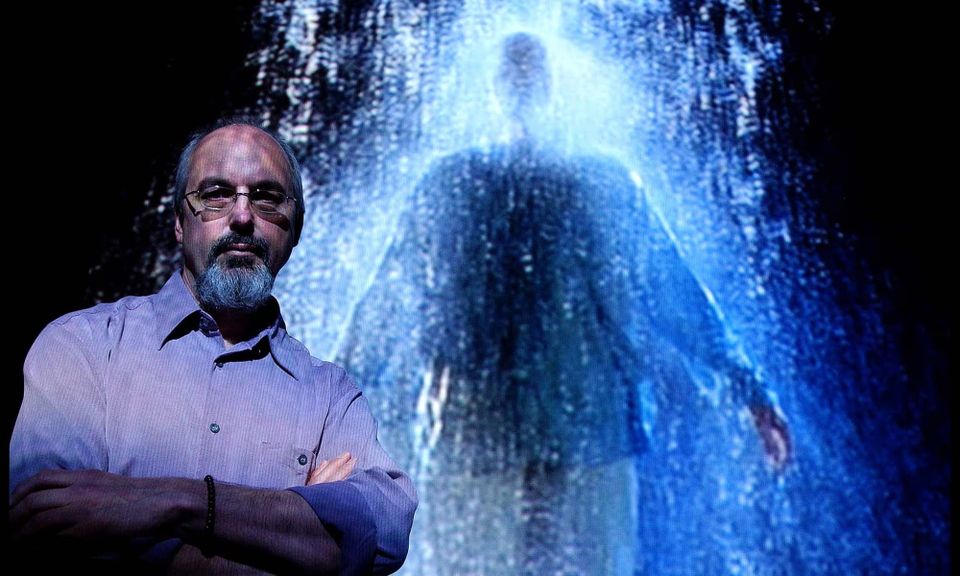 A balding man in glasses in a button-down shirt stands in front of a pixilated video image of a figure with water splashing down on it.