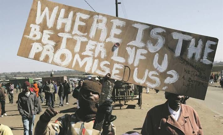 African protestors grouped around a masked man holding a wooden sign that reads WHERE IS THE BETTER LYF U PROMISED US?