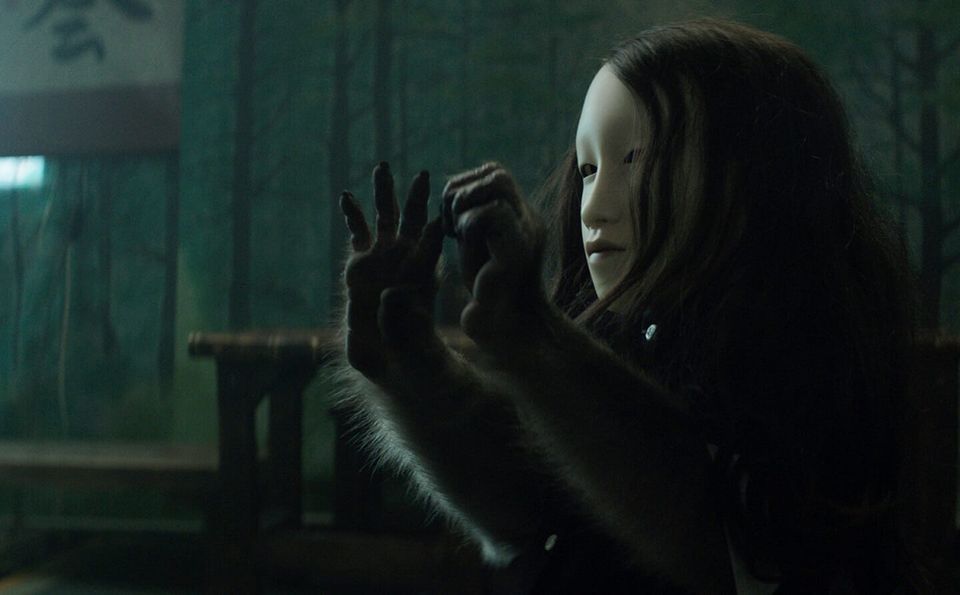 A woman with long dark hair in a white mask with the arms and hands of an animal.