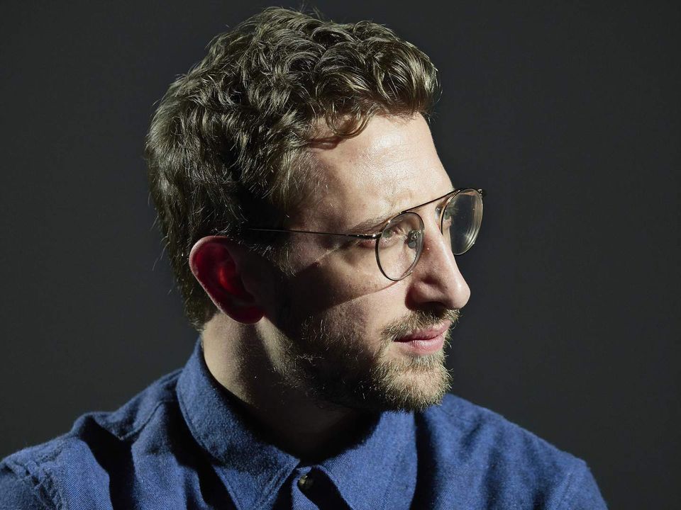 A man in profile, with curly blonde hair, short beard, glasses and wearing a collared blue shirt.