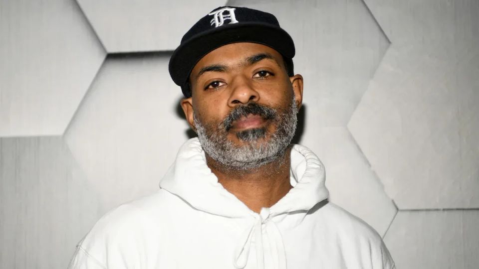 A Black man with a gray beard wearing a dark-colored Detroit Tigers baseball cap and a white hooded sweatshirt.