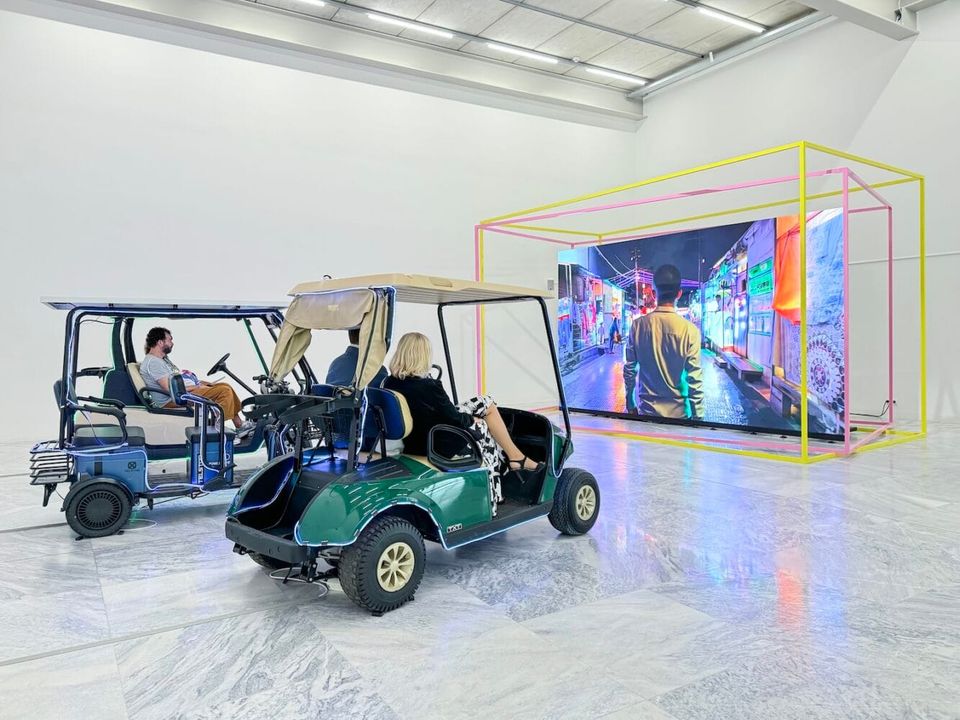 A gallery with a large screen framed in yellow and pink bars, with two golf carts positioned in front with exhibition visitors sitting inside and watching the video on screen.