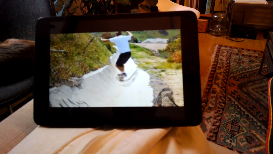 A tablet with an image of a skateboarder, positioned on a couch in a living room with a patterned carpet in view.