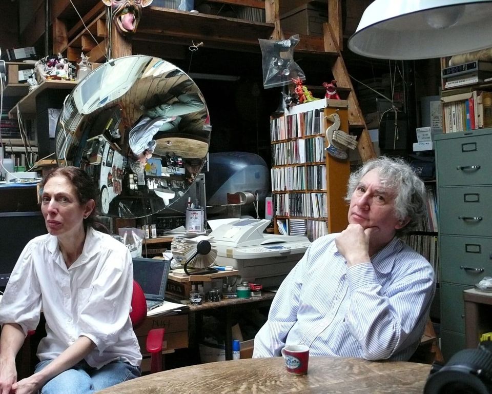 A man and a woman sitting in a work space filled with books, papers and ephemera.
