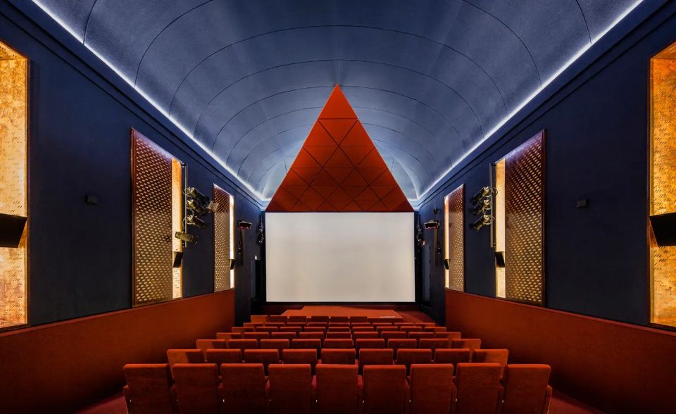 The interior of a cinema with a large red triangle above the screen, red plush seats, a blue ceiling, and golden wall panels.