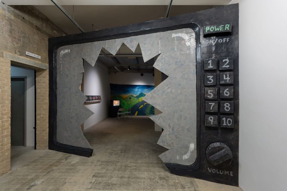 A gallery view of an immense television with a broken screen that visitors walk through to access another gallery with monitors and tapestries.
