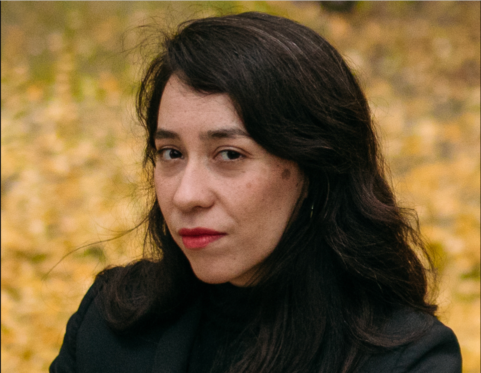 A woman wearing black with long black hair looking at the camera, posing against an out of focus autumn background.