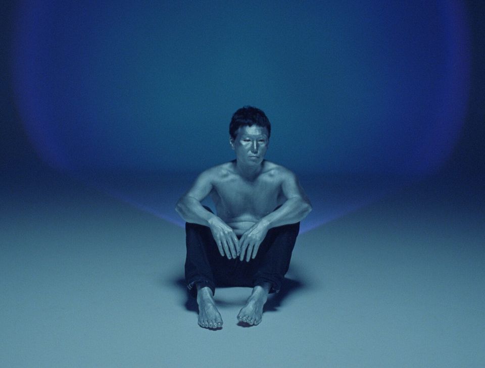 A man wearing black pants and no shirt sits against a nondescript turquoise backdrop, his gaze focused on something beyond the camera.