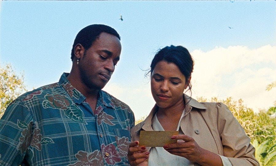 A Black man and a Black woman stand in a lush green landscape, reading a postcard.