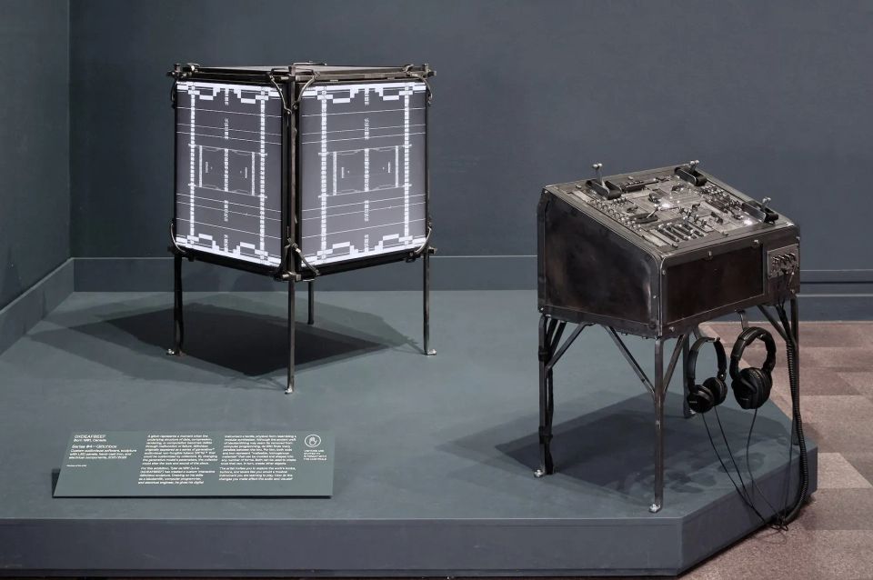 A four-sided screen device showing digital art on a gallery platform with a control panel next to it with various switches, levers and headphones.