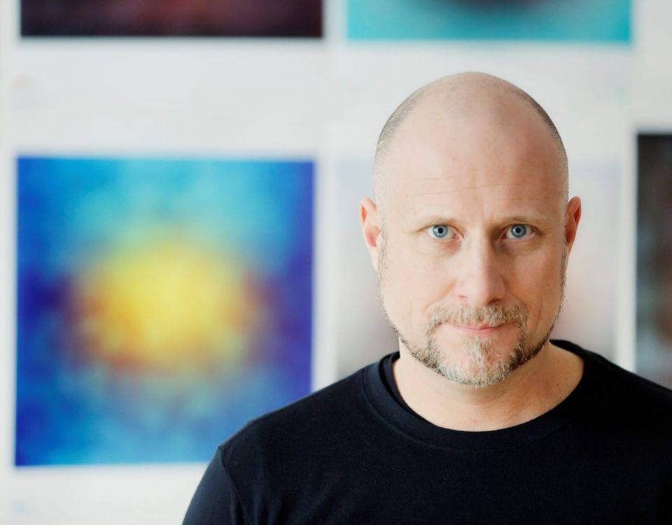 A balding man with blue eyes and a thin beard, wearing a black shirt, standing in front of a painting that is out of focus in the distance.