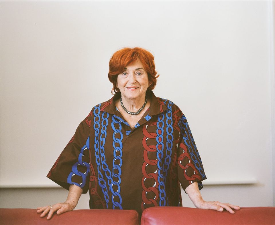 A smiling older woman with red hair, standing behind a maroon couch, wearing a brown short with multicolored designs of chains.