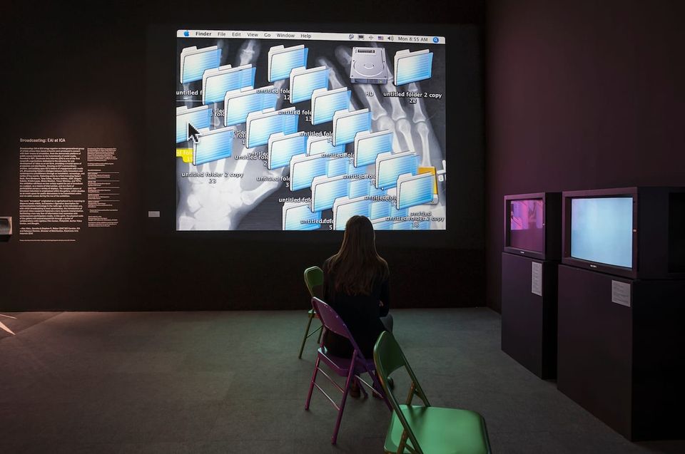A woman sits on a chair in a dark gallery in front of two monitors, with a large projected image of a computer desktop screen in the background.