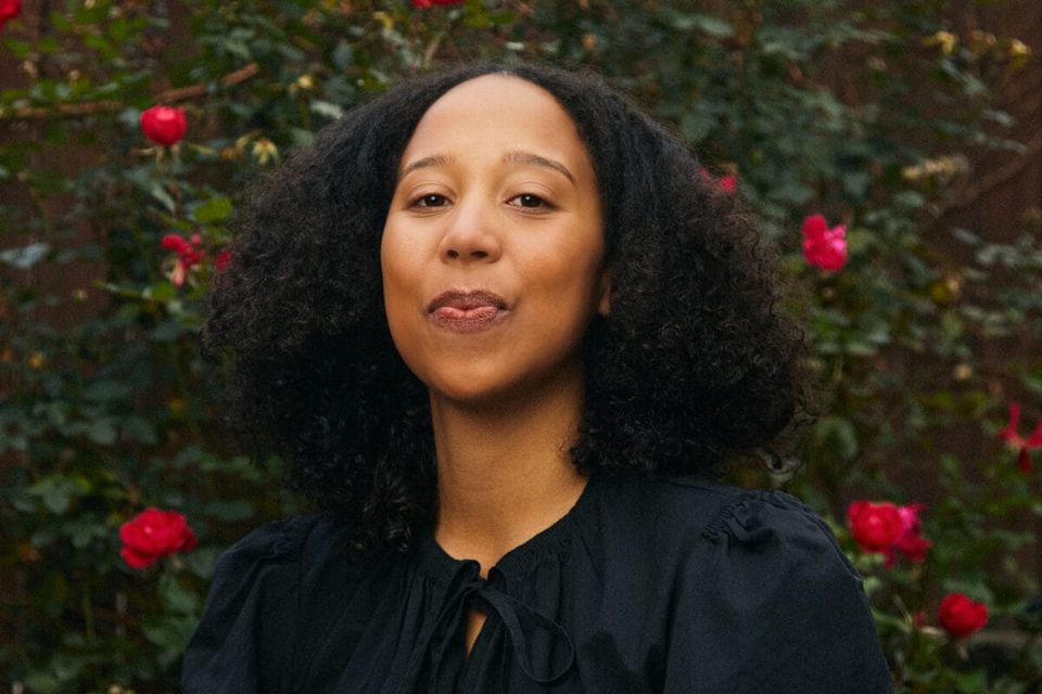 A Black woman with long hair, wearing a black dress, standing in front of a bush with red roses.